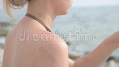 一个迷人的年轻女孩在阳光明媚的夏天坐在海滨，在她的肩膀上涂上防晒霜。 夏季